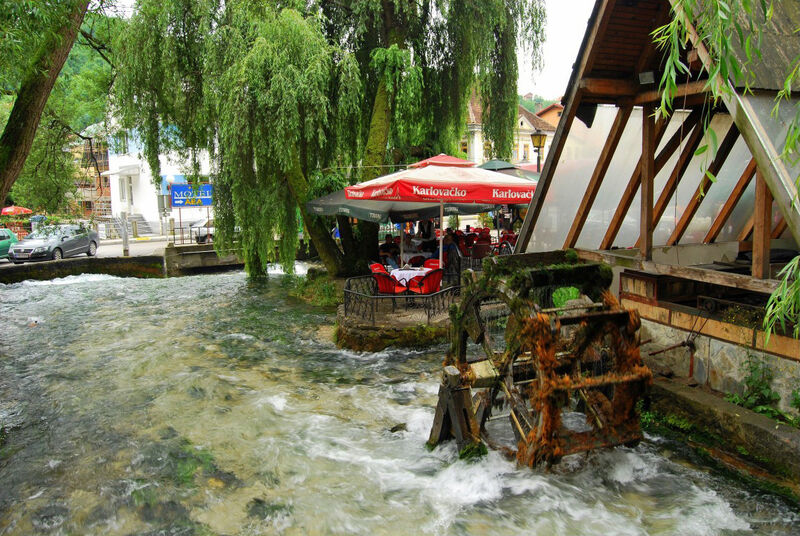 A wooden water wheel in a flowing river next to a restaurant outdoor seating area under green trees.