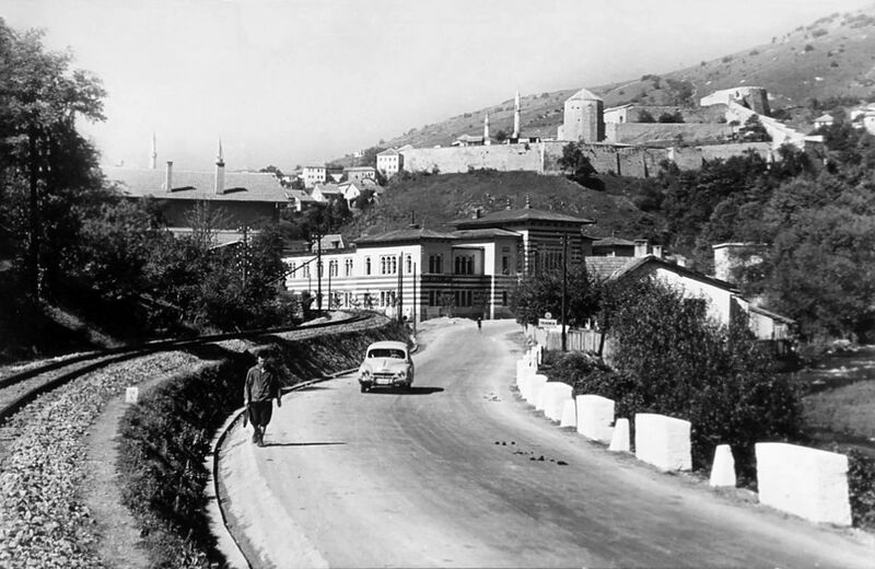 Black and white view of a road with a vintage car and person leading toward a town with a fortress on the hill.