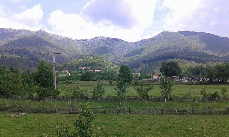 A wide landscape view of green fields, a wooden fence, and mountains under a cloudy sky.