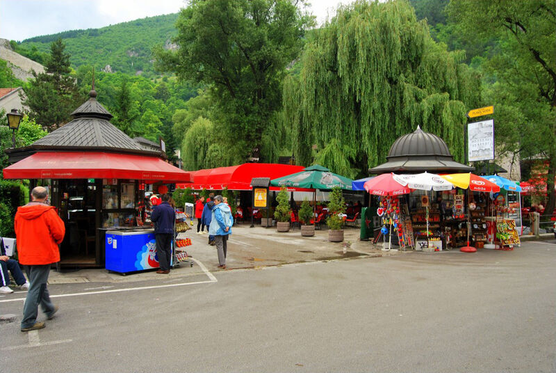 A street scene in Travnik featuring small kiosks, souvenir shops, and outdoor cafes surrounded by green trees.