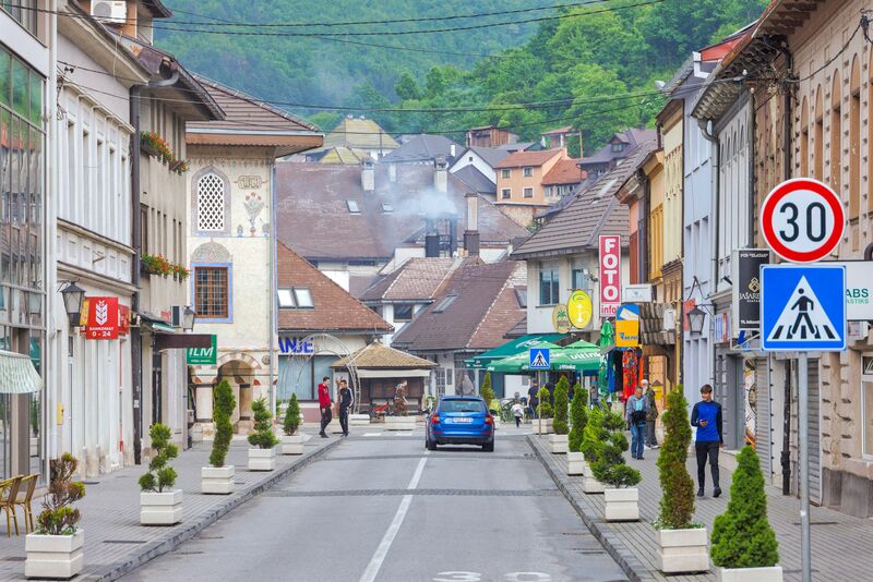 A street view in Travnik featuring the decorated exterior of the Šarena Mosque and surrounding shops.